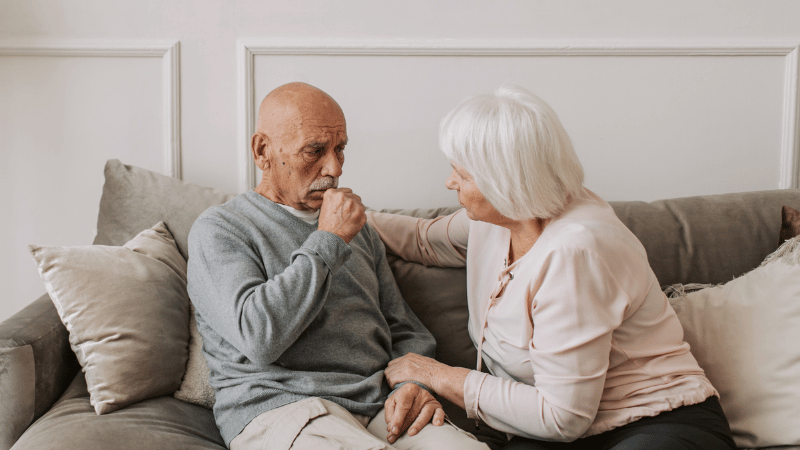 Elderly man coughing while an elderly woman looks at him with concern