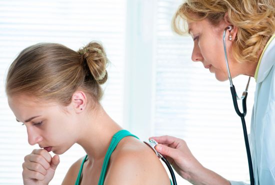 Teenage girl coughing while a clinician holds a stethoscope against her back