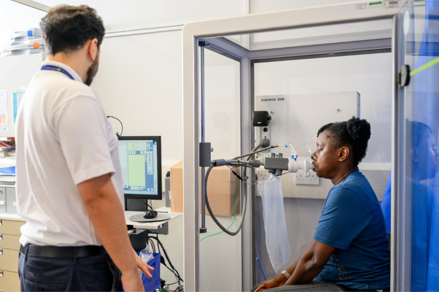 Patient performing a lung function test inside a clear body plethysmography booth while a clinician observes
